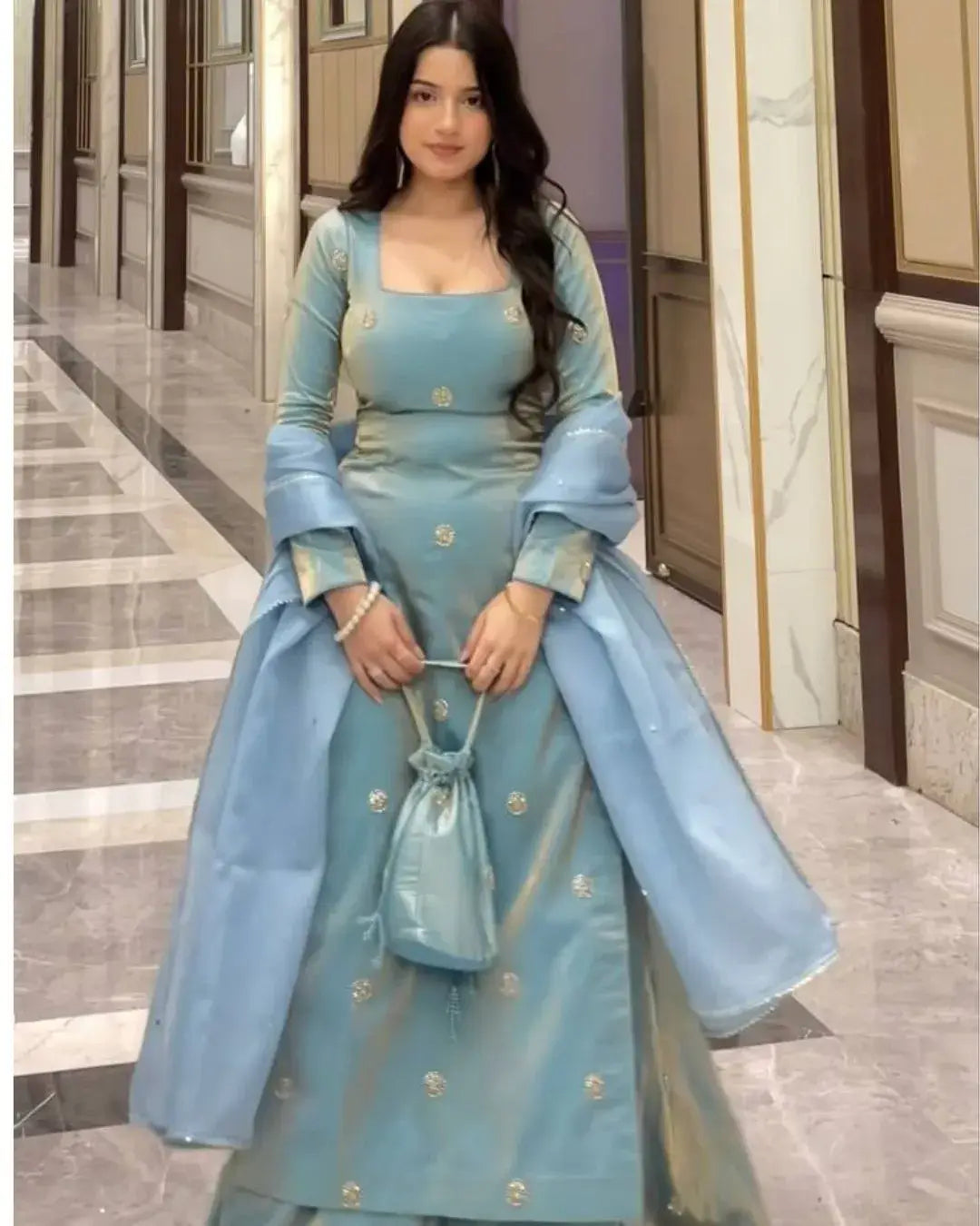 Woman in a light blue traditional outfit holding a matching bag in a hallway.
