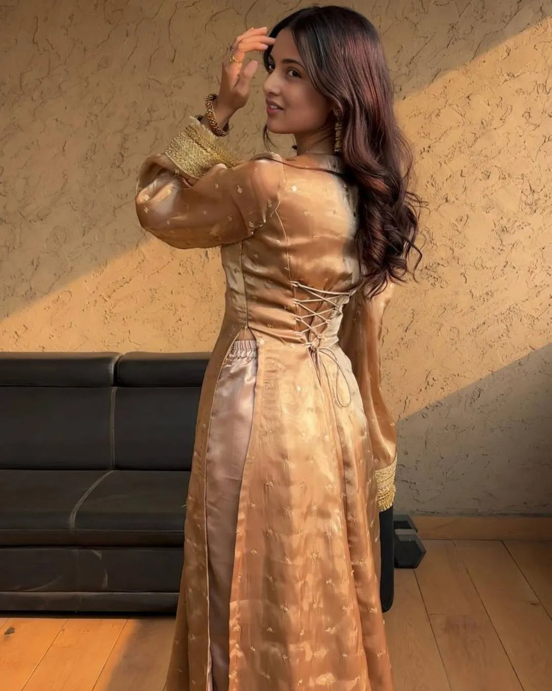Woman in a gold dress standing in a room with a textured wall and black sofa.