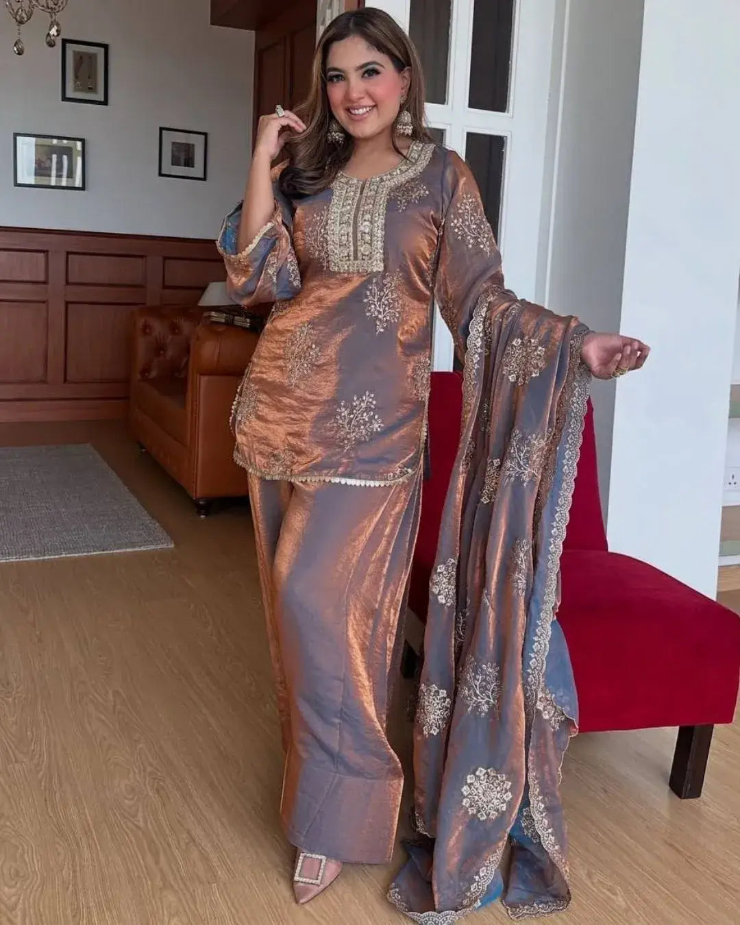 Woman in a traditional outfit standing in a room with wooden paneling and a red chair.