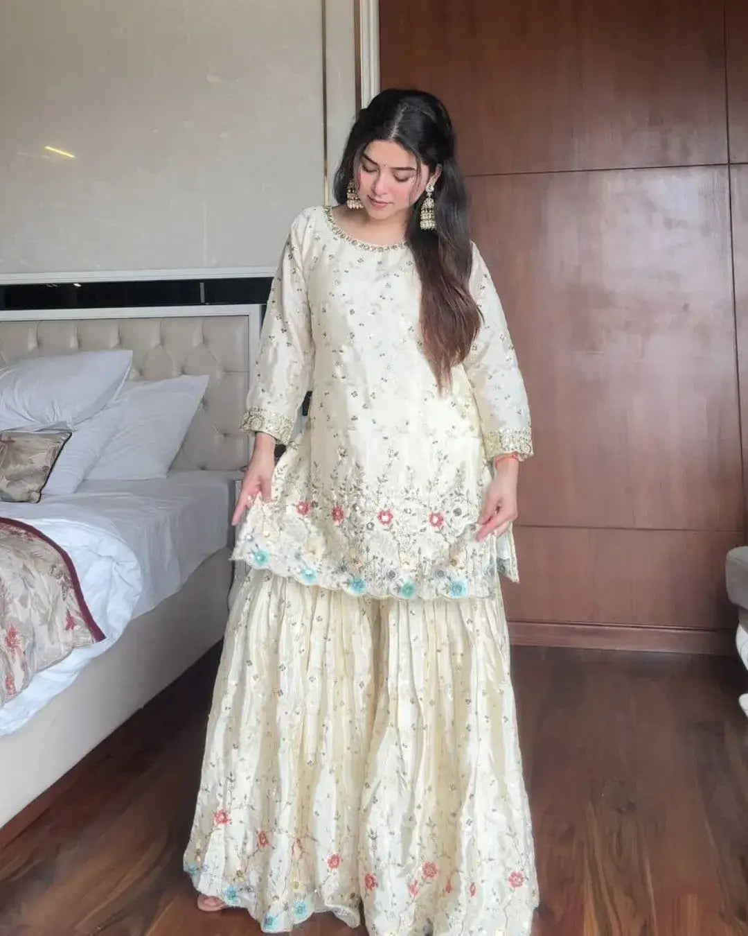 Woman in a light-colored traditional outfit standing in a bedroom.