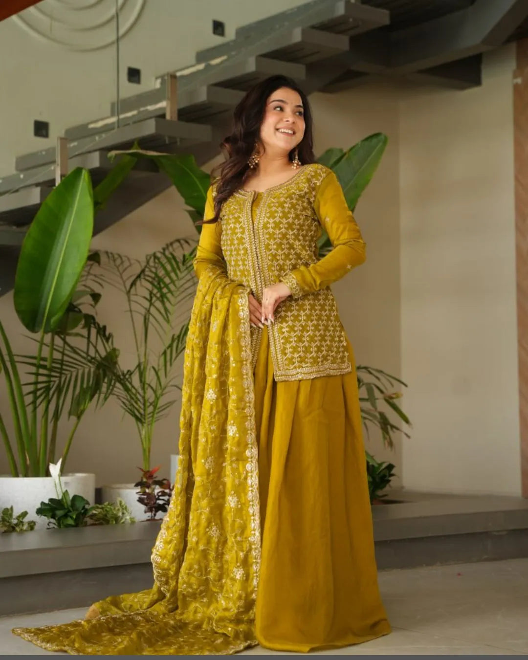 Woman in a yellow sharara outfit standing indoors with plants in the background