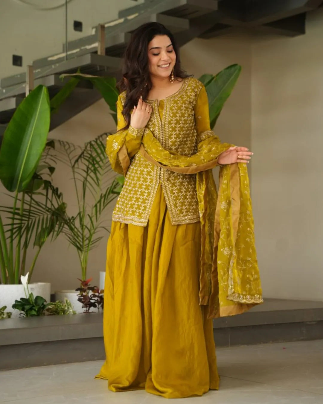 Woman in a yellow sharara outfit standing indoors with plants in the background