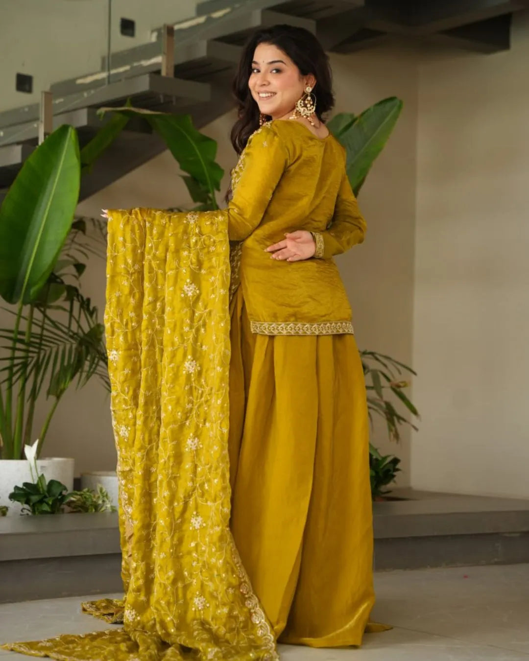 Woman in a yellow traditional outfit with a matching dupatta, standing indoors with plants in the background.