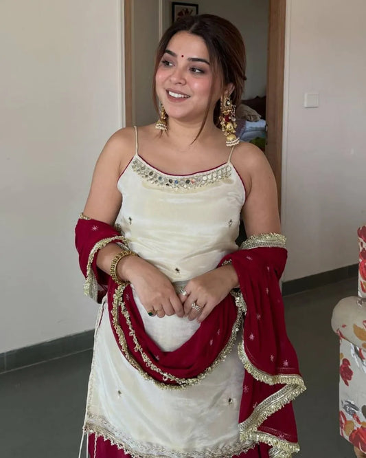 A woman wearing a traditional South Asian outfit with an off-white embroidered top and a deep red dupatta draped over her arms, standing in an indoor setting.