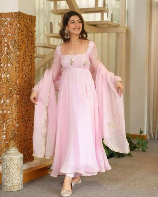 A woman models a light pink traditional Indian anarkali gown with matching sheer dupatta and silver embellishments, set in an indoor environment with a wooden staircase and carved screen.