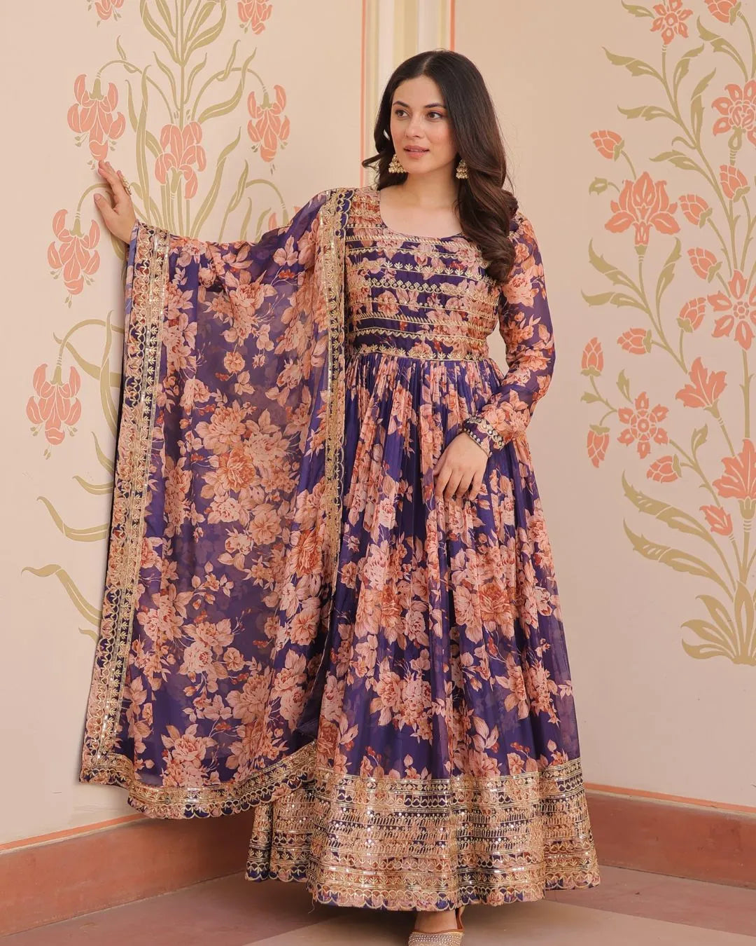 Woman wearing a floral dress with a matching dupatta against a decorative wall.