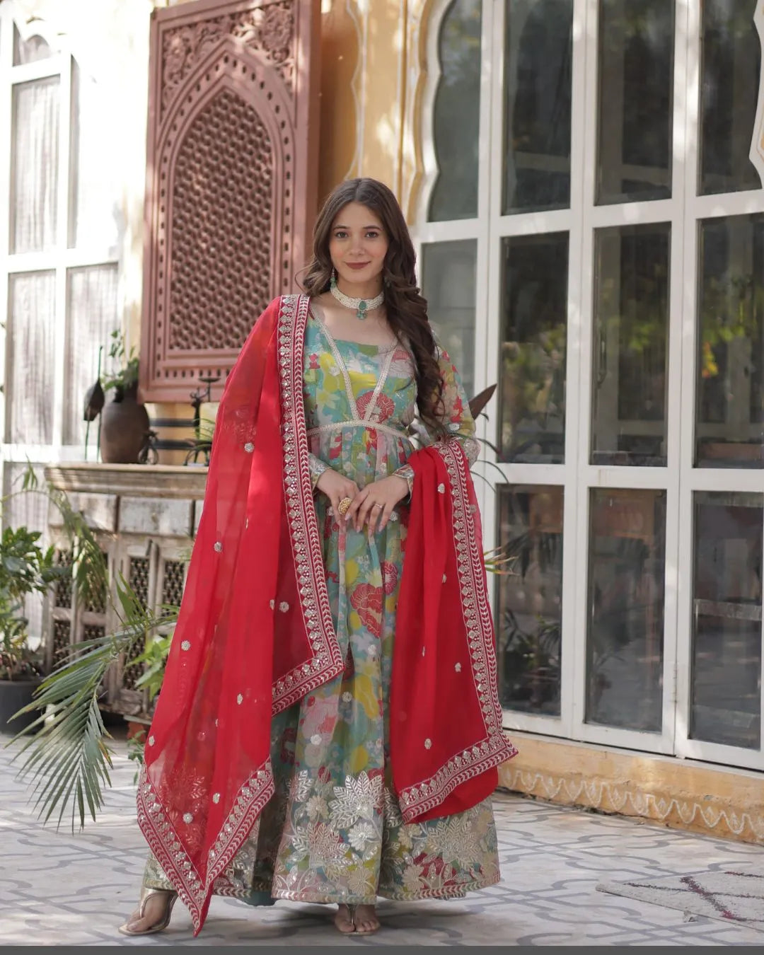 Woman in traditional outfit with red dupatta standing in front of decorative wall.