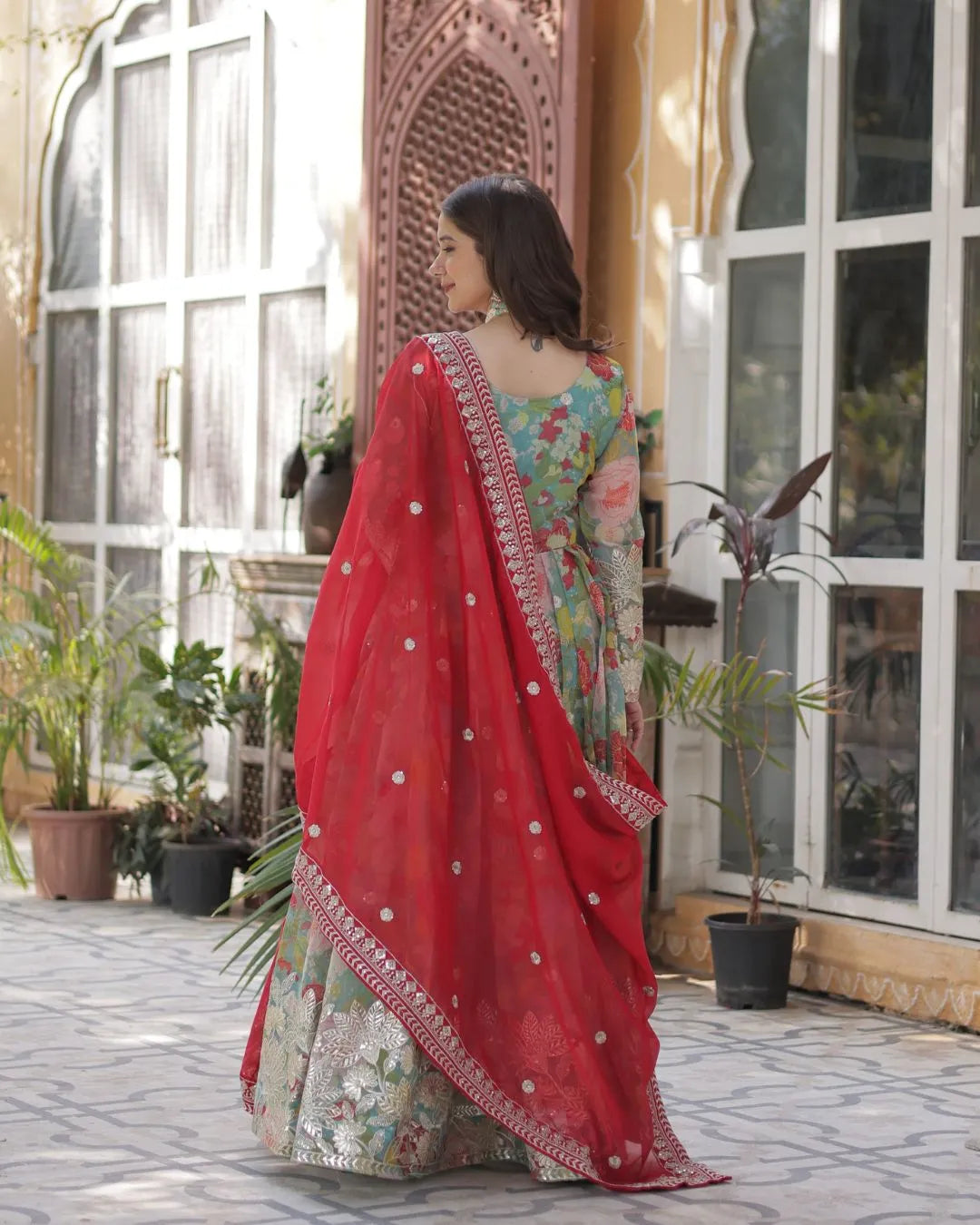 Woman in a floral dress with a red dupatta standing in front of a decorative archway.