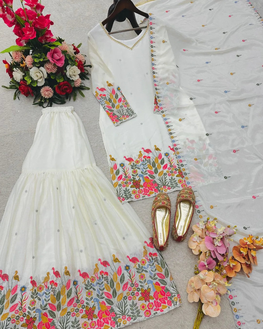 White embroidered outfit with colorful patterns, gold shoes, and flowers on a light gray background