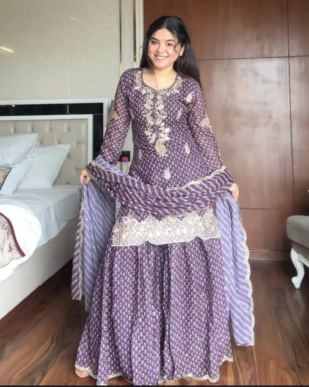 Woman holding a purple traditional outfit with white patterns in a bedroom setting