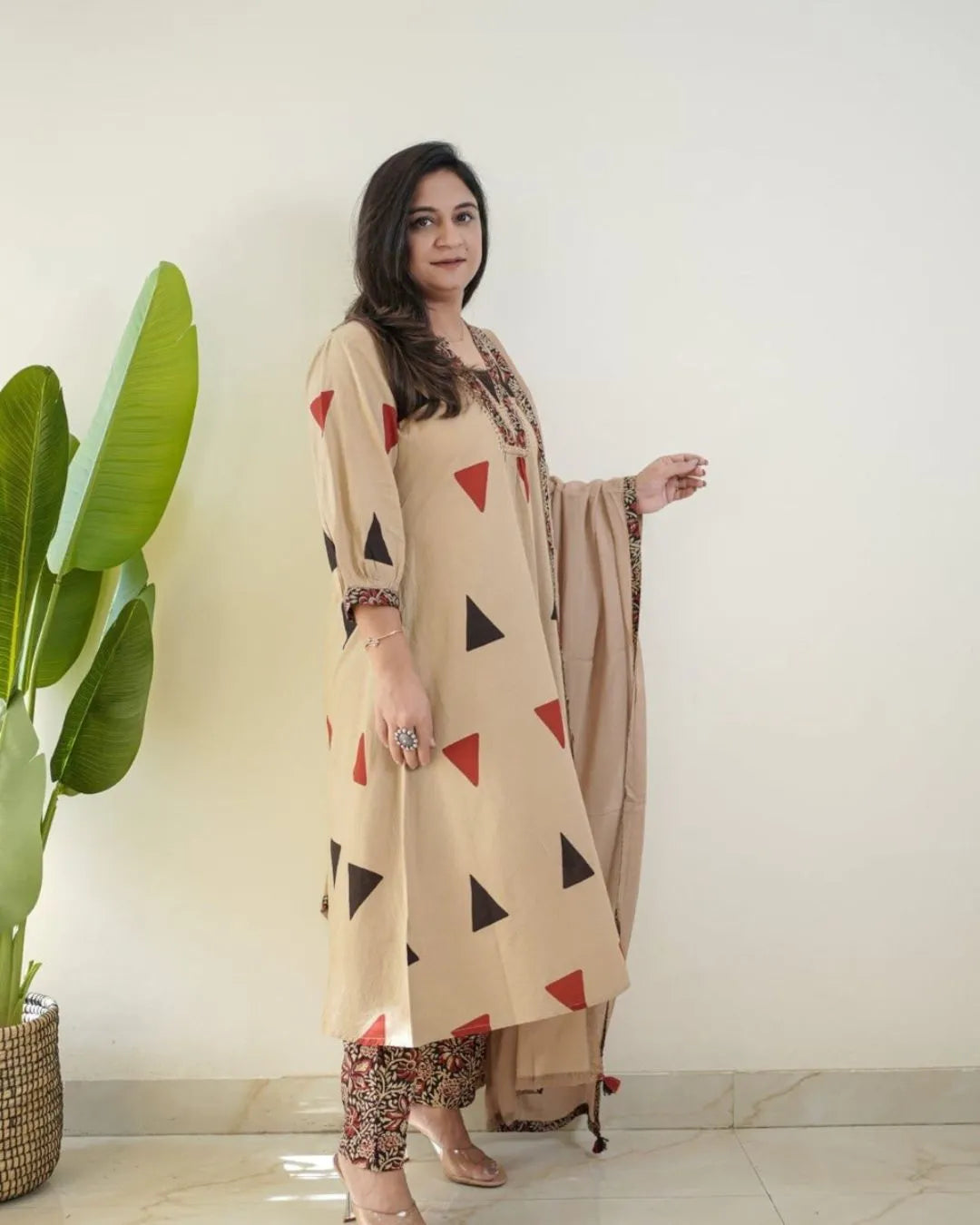Woman wearing a beige top with red triangle patterns and floral embroidery, standing against a white wall.