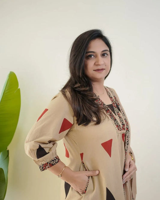 Woman wearing a beige top with red triangle patterns and floral embroidery, standing against a white wall.