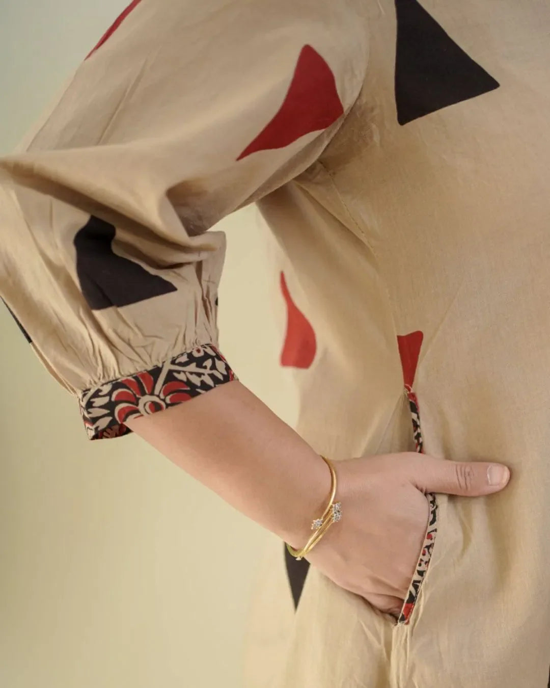 Woman wearing a beige top with red triangle patterns and floral embroidery, standing against a white wall.