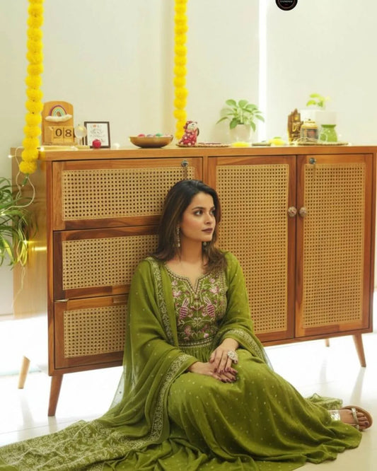 Woman in a Green Colour Anarkali Suit Set sitting in front of a wooden cabinet with decorative items.