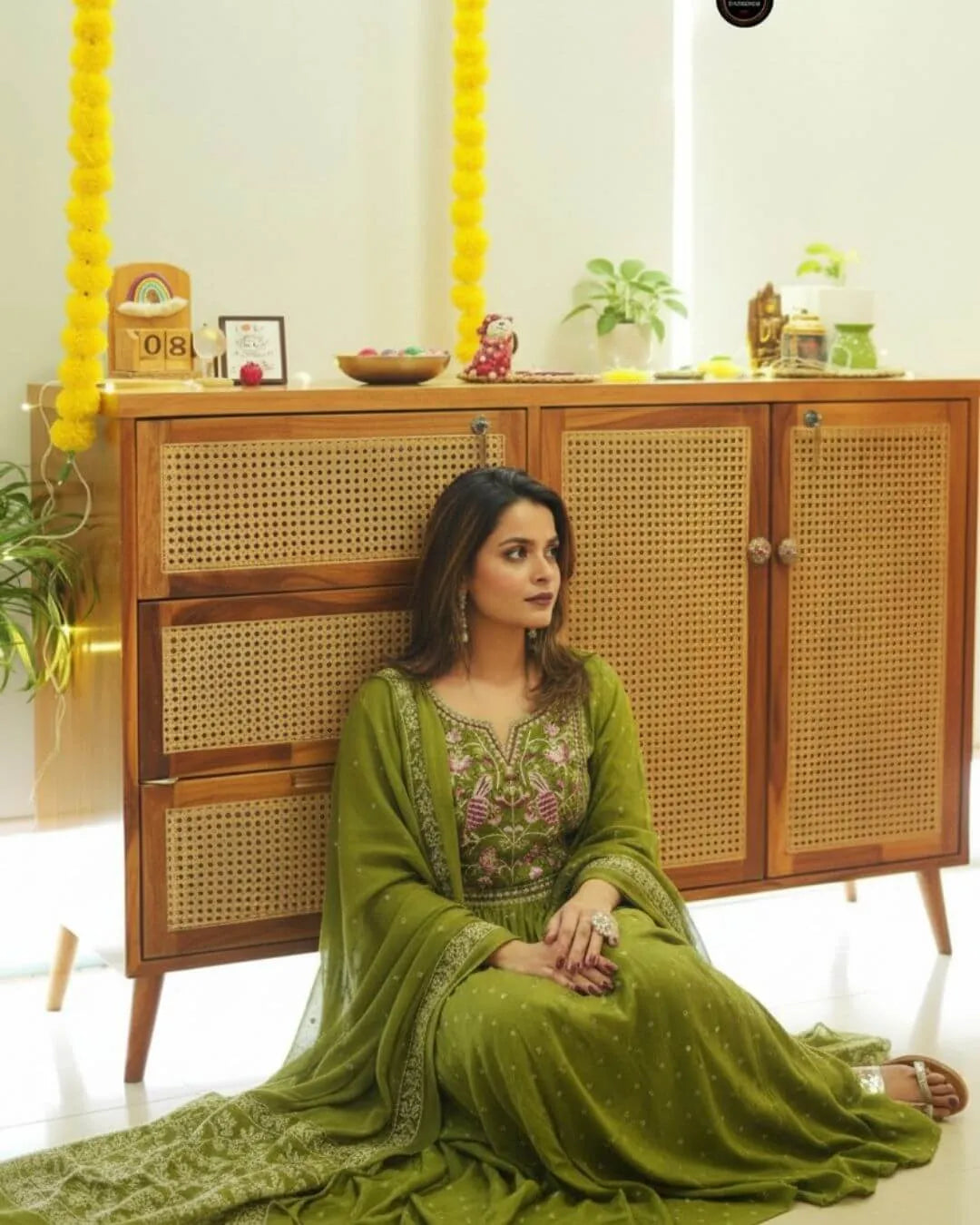Woman in a Green Colour Anarkali Suit Set sitting in front of a wooden cabinet with decorative items.
