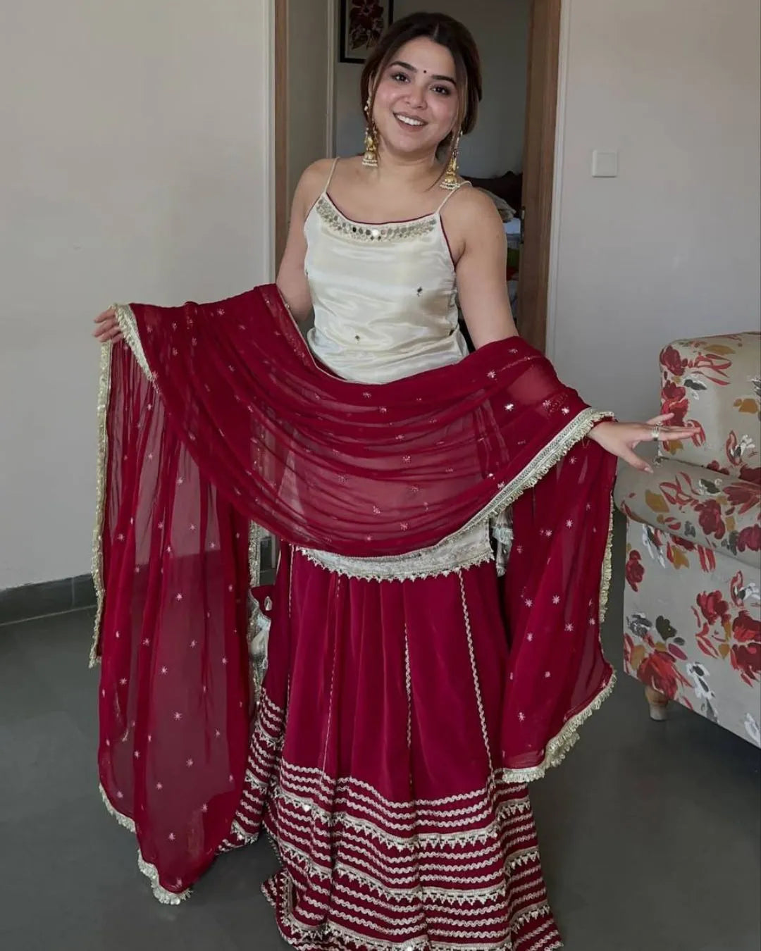 A woman modeling a traditional Indian outfit consisting of a deep red skirt with white embroidery, a matching red dupatta, and a cream-colored sleeveless top, standing in an indoor setting.