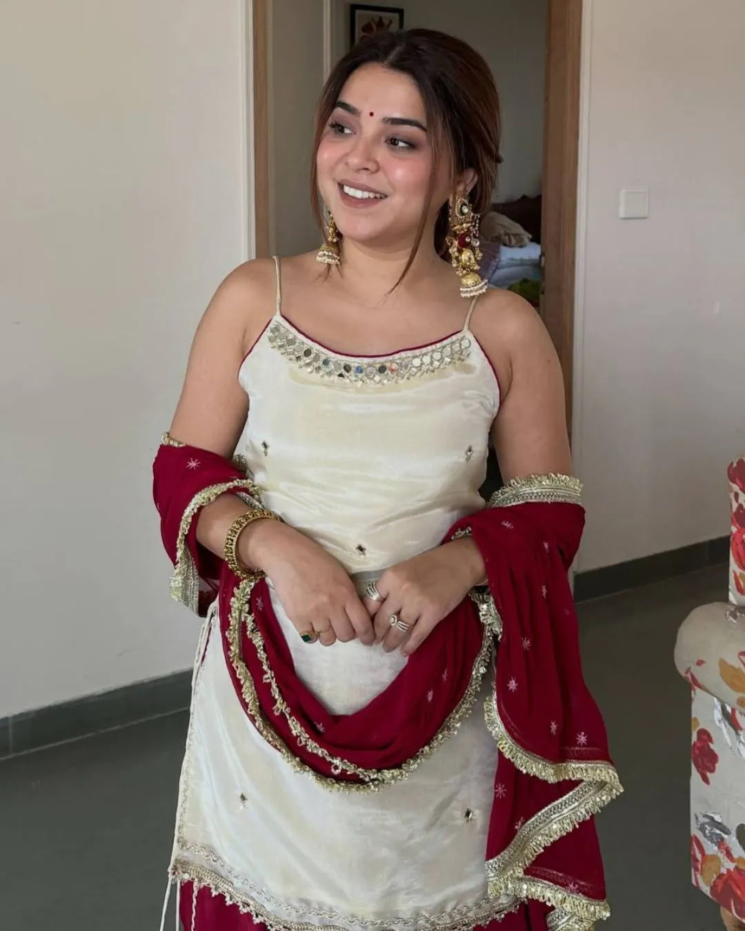 A woman wearing a traditional South Asian outfit with an off-white embroidered top and a deep red dupatta draped over her arms, standing in an indoor setting.