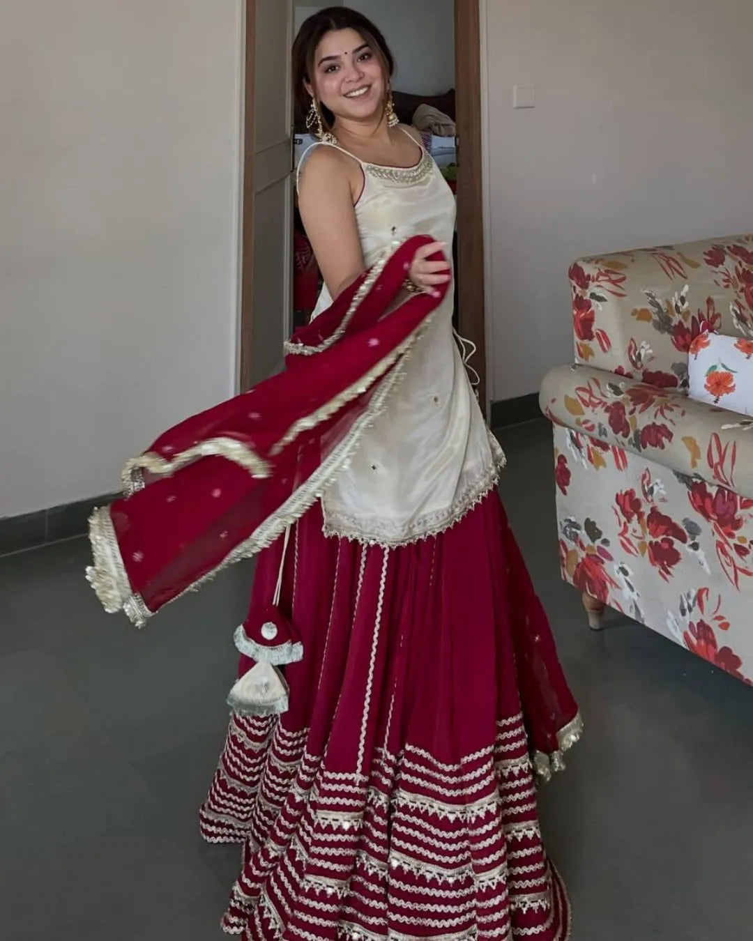 A woman modeling a traditional Indian outfit consisting of a cream sleeveless top, a deep red flared skirt with white embroidery, and a matching deep red dupatta with gold borders, standing in an indoor room.