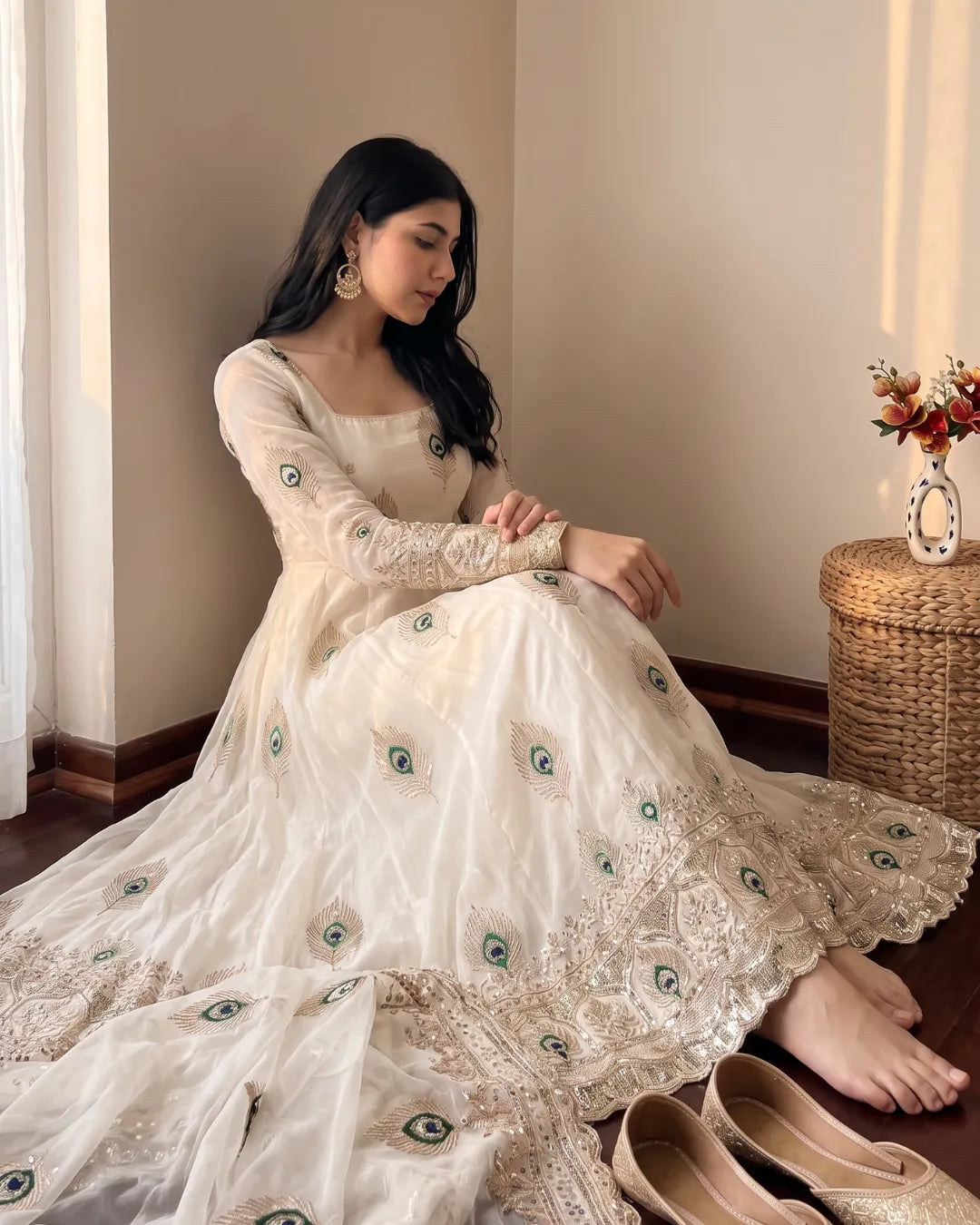 A woman sits barefoot on a wooden floor wearing a long-sleeved cream/off-white embellished gown with peacock-eye embroidery suit set. She is posed in a warm, sunlit room with a woven side table and flowers in the background.