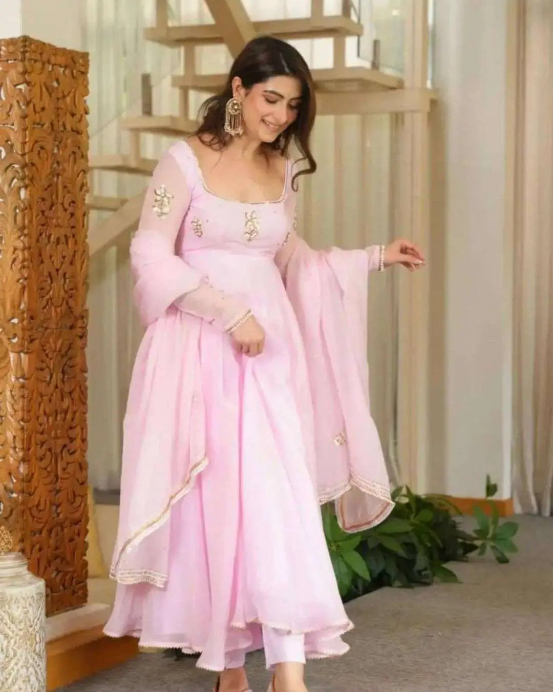 A woman wearing a light pink traditional outfit with gold embroidery, standing in an indoor setting with a staircase and carved wooden panel in the background.
