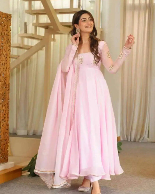 A woman modeling a light pink traditional Anarkali gown with long sleeves, a matching draped scarf, and gold embroidery on the sleeves and border, set in an indoor environment with a staircase and curtains.