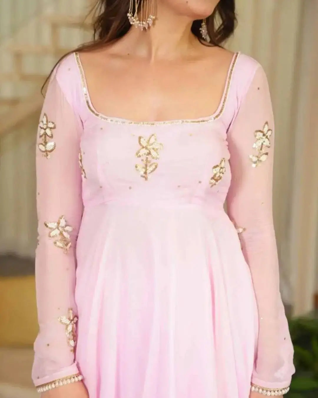 A woman wearing a light pink traditional dress with sheer long sleeves, gold floral embroidery, and a square neckline.