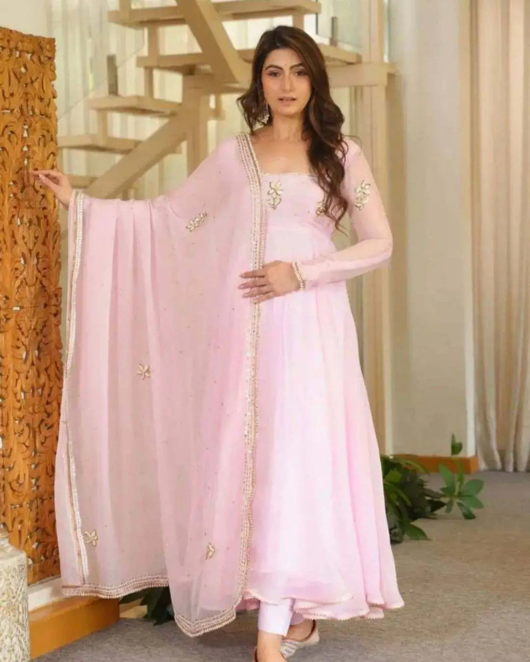 A woman modeling a light pink traditional Indian outfit with a matching long dupatta, featuring gold embroidery, in an indoor setting with a staircase and wooden screen.