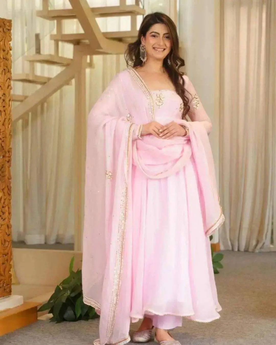 A woman modeling a light pink traditional Indian outfit with a matching dupatta, featuring gold embroidery on the neckline and sleeve borders, set against a background of cream curtains and wooden stairs.