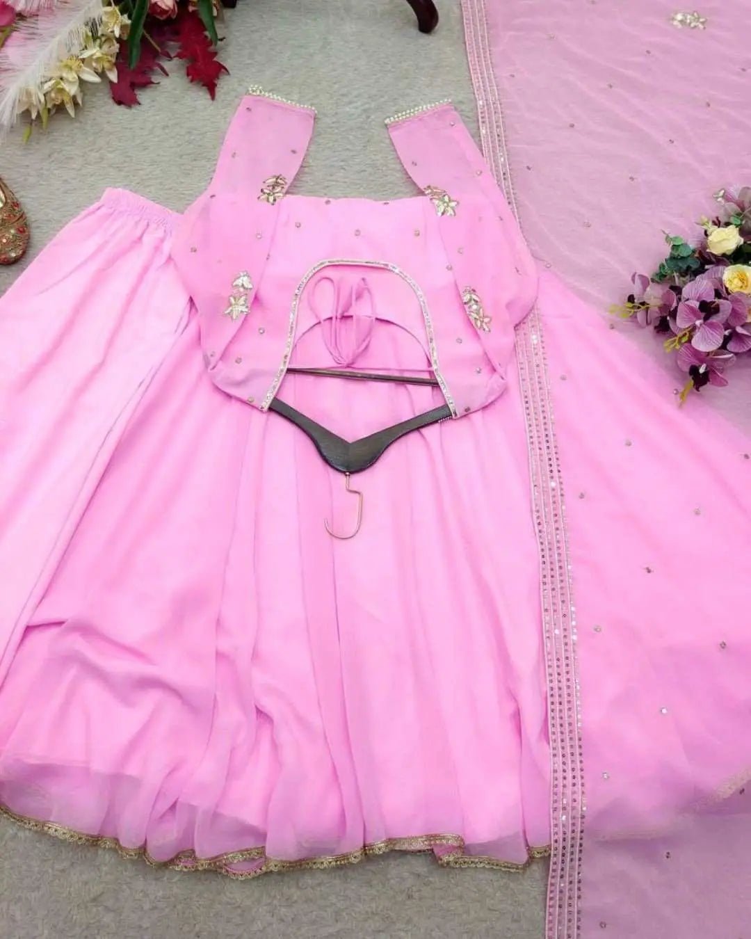 A pink traditional dress set laid flat on a gray surface, featuring a pink kurta with gold embellishments, a matching flared skirt, and a sheer pink dupatta with sequined borders.