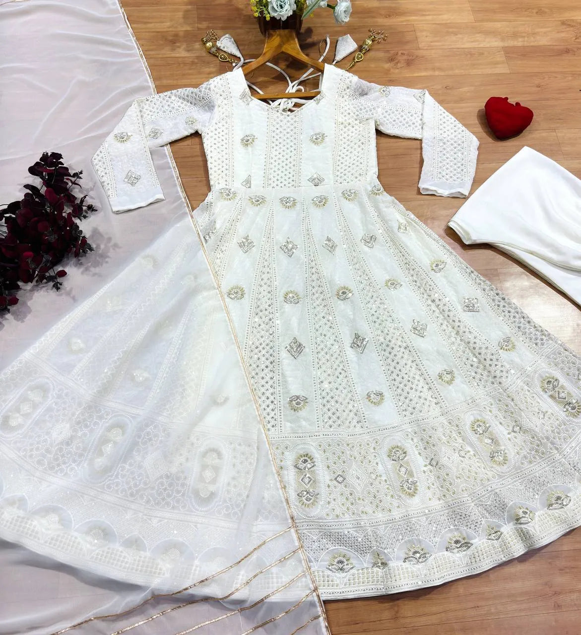 A white embroidered long-sleeve dress with a matching sheer dupatta, laid flat on a wooden floor with decorative props including a small bouquet, a red heart-shaped box, and folded white fabric.