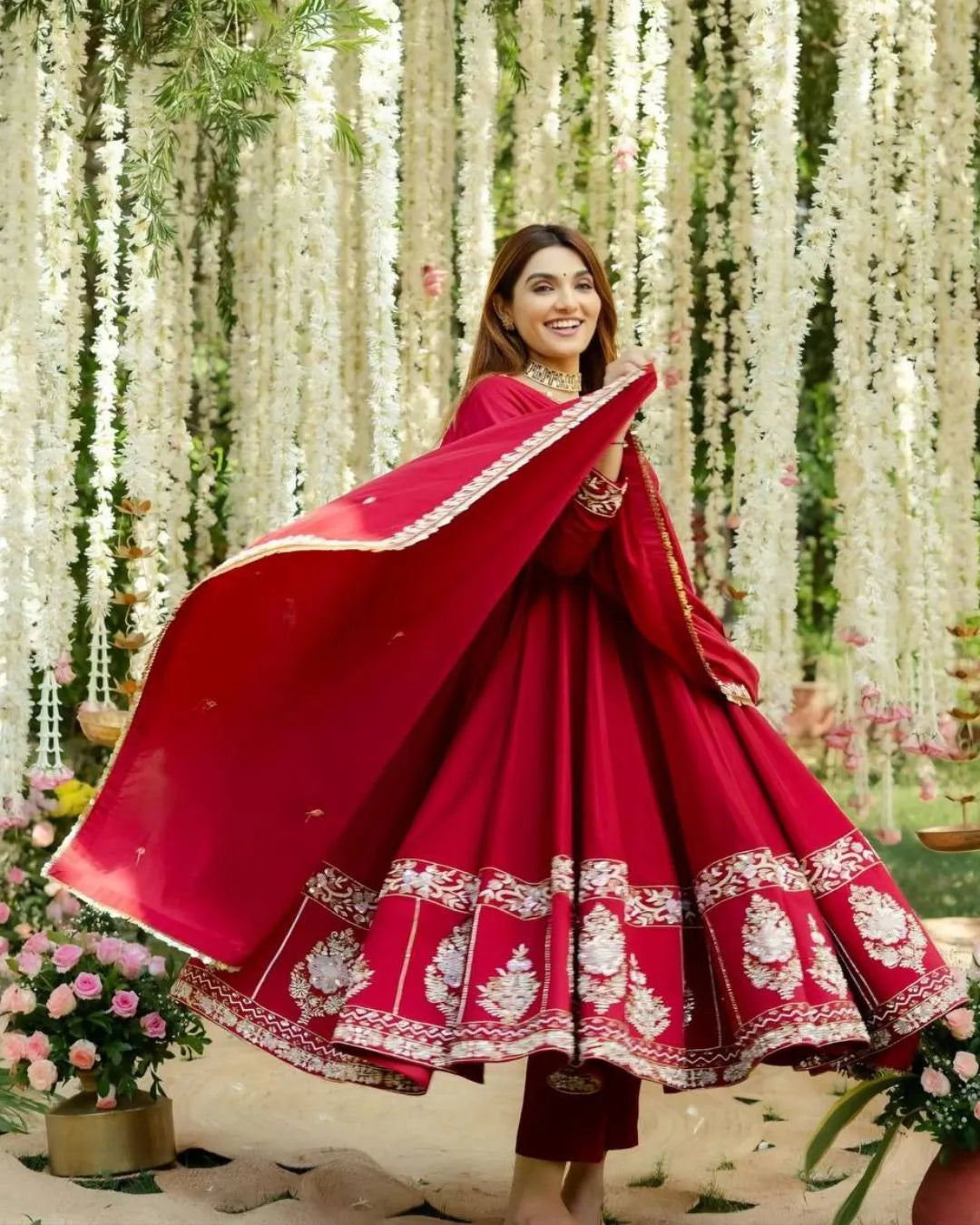 Woman in a red traditional outfit with white embroidery standing amidst floral decorations.