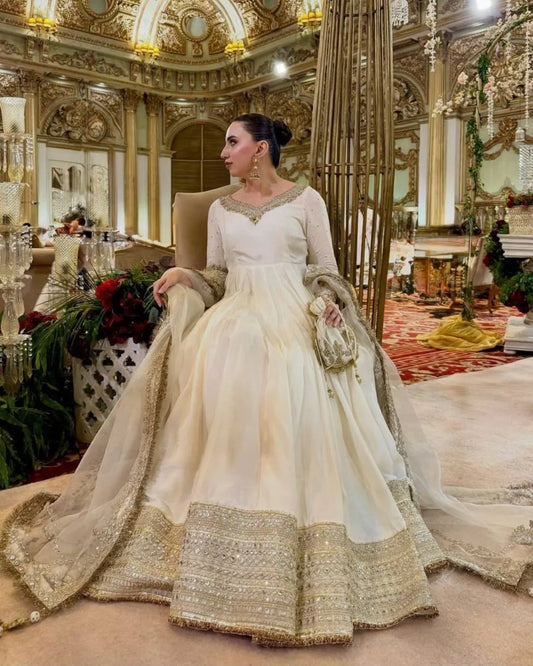 A woman wearing an ivory and gold heavily embellished traditional Anarkali gown with a matching dupatta, standing in an ornate, luxurious event hall.